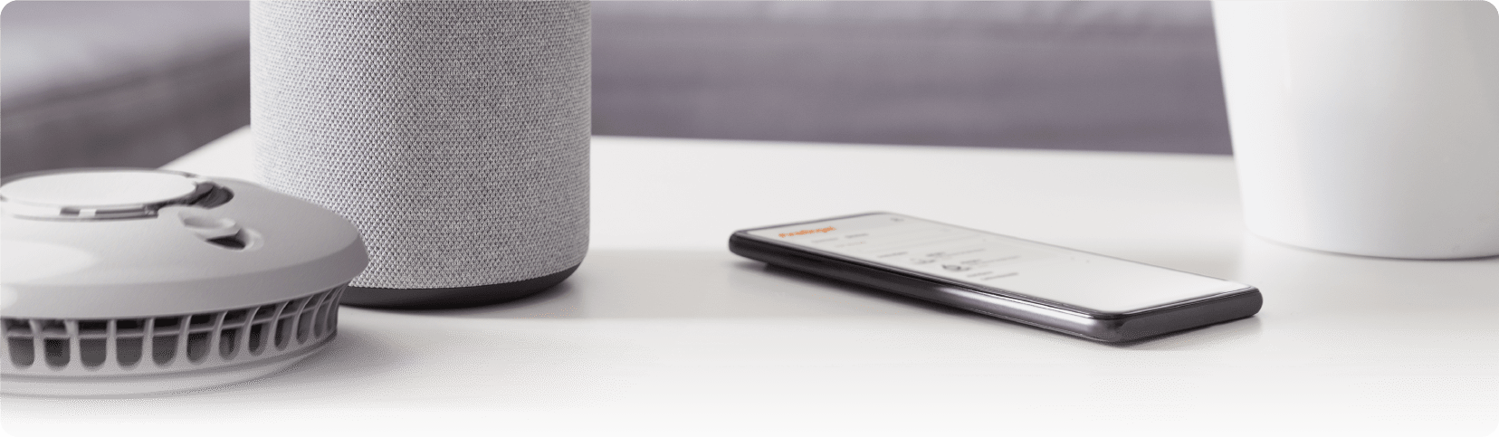 A smoke detector, smart speaker, and smartphone for Dementia Action Week on a white table.