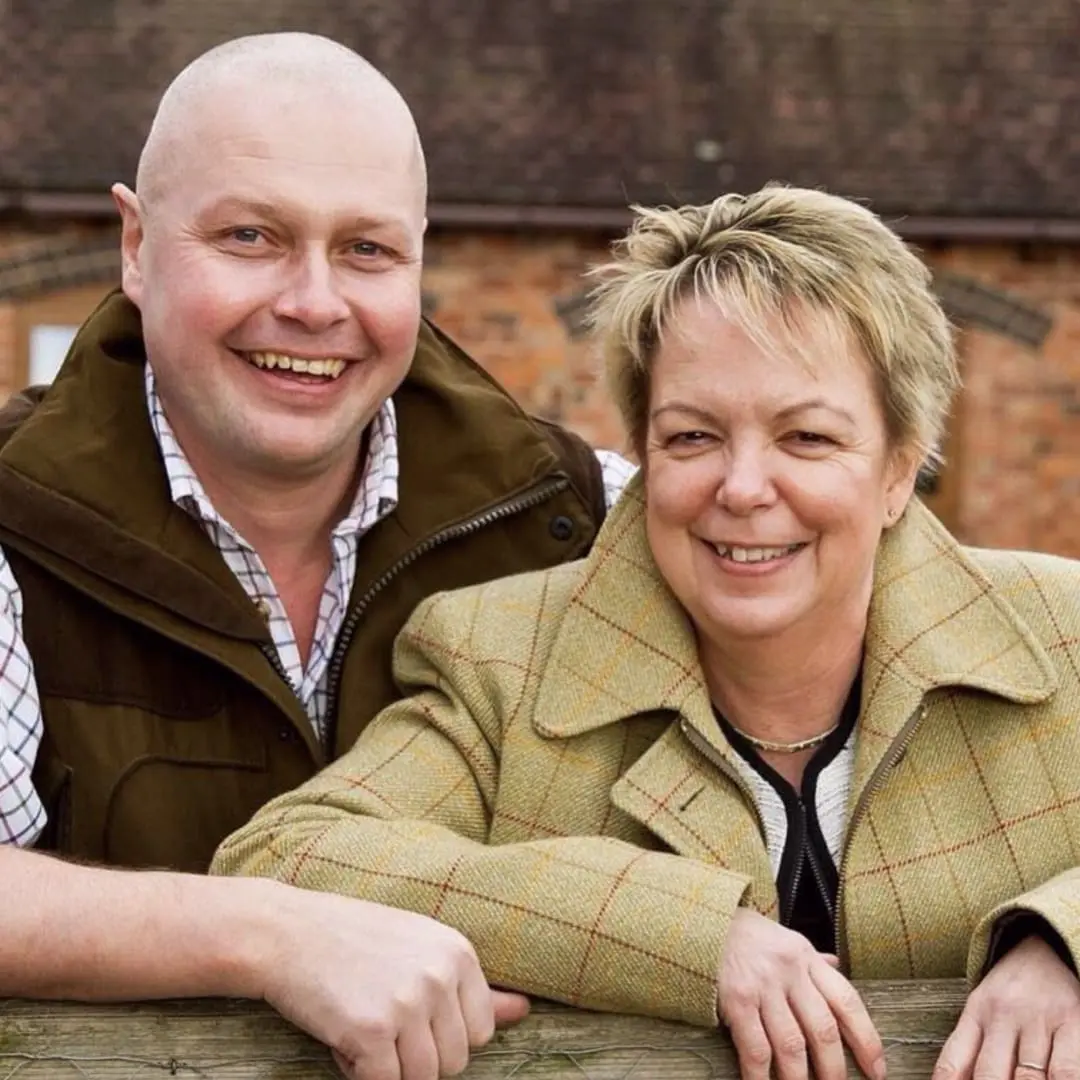 Two smiling adults at a family farm stay, leaning on a fence with a brick building behind them.