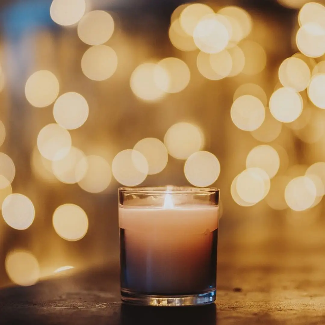 A lit candle in a glass holder with warm, blurry lights and festive fire tips in the background.