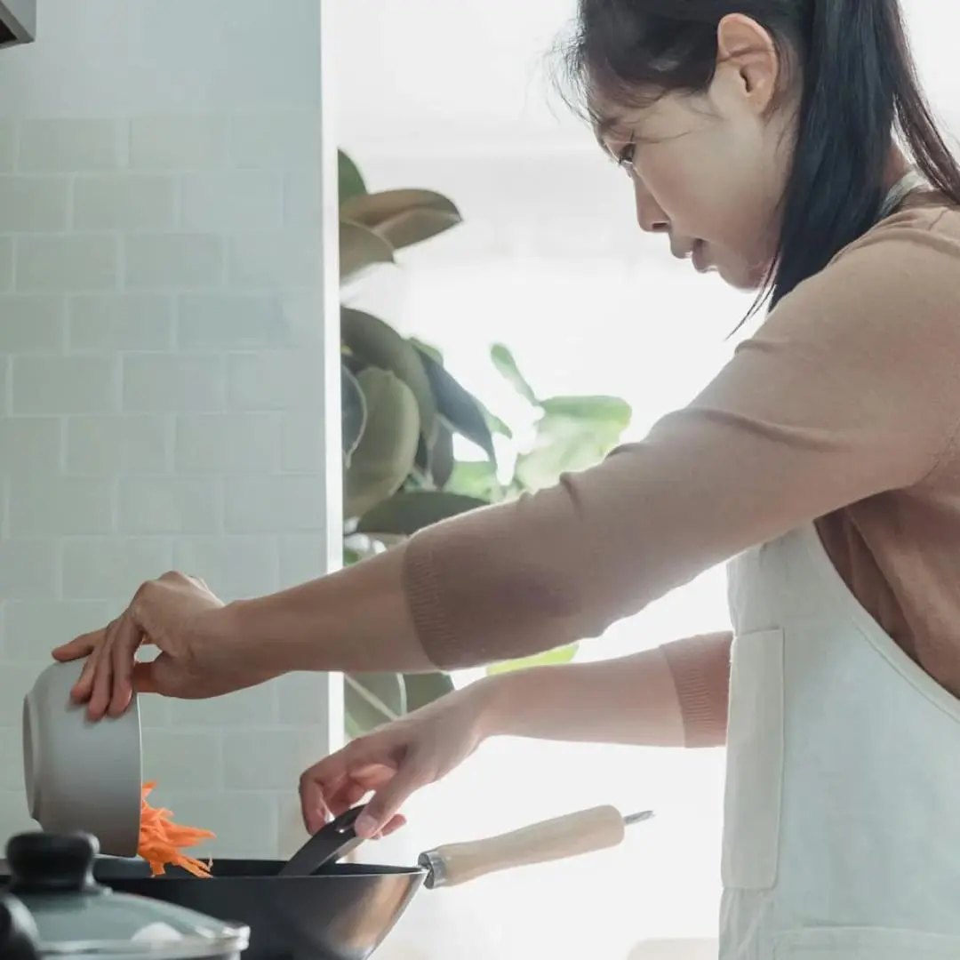 Woman in apron adds carrots to a frying pan, demonstrating good kitchen safety.