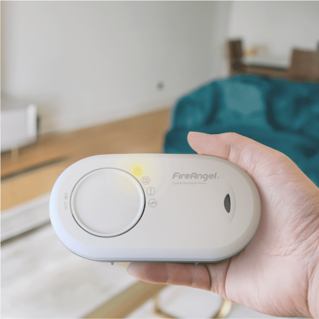 Hand holding a FireAngel carbon monoxide alarm in a modern living room setting.
