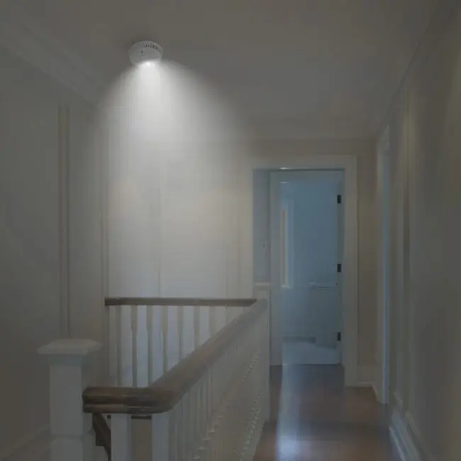 The FA6611-R smoke detector emits light on a hallway ceiling near the stairs and open door.
