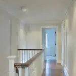 Bright corridor with white walls, wooden handrail, and FA6615-R doorway leading to a bathroom.