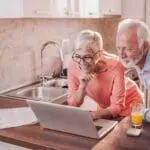 Smiling older couple in kitchen view FP1000W2-R info on laptop with drinks and papers.
