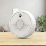 An SB1-R smoke detector sits on a wooden table with a blurred lounge in the background.