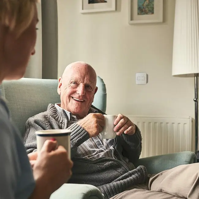 Older man smiling in armchair with mug, chatting, in FS1524W2-T jumper.