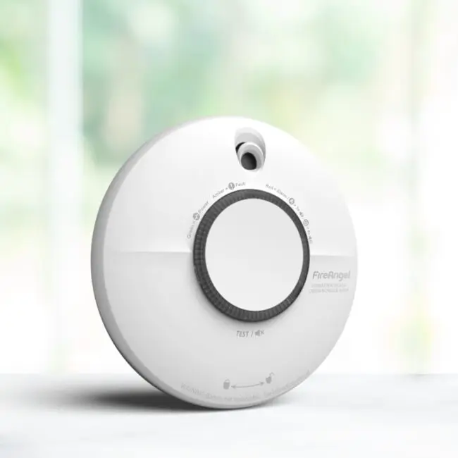 A white SCB10-R smoke and CO alarm on a light surface with a blurred background.