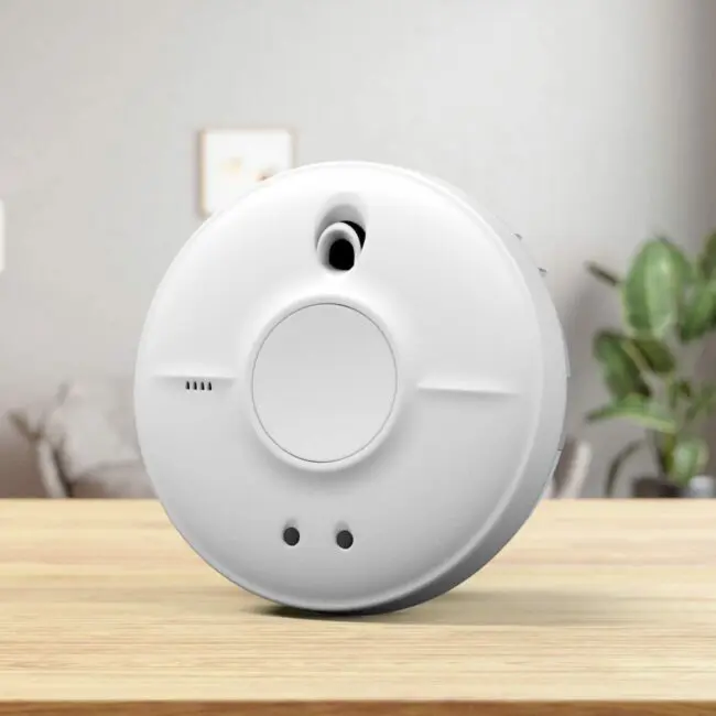 The SW10-EUT smoke detector sits on a wooden table with a blurred lounge background.