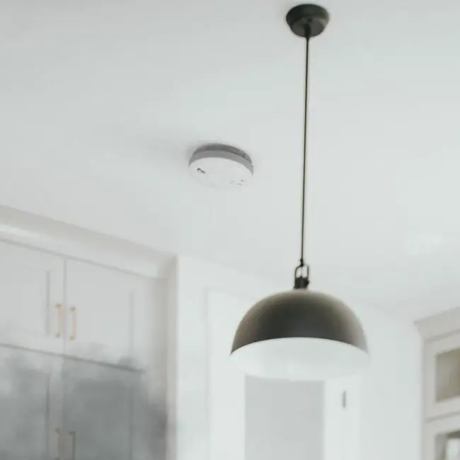 An SW1-R smoke detector with a black pendant light on a white kitchen ceiling.