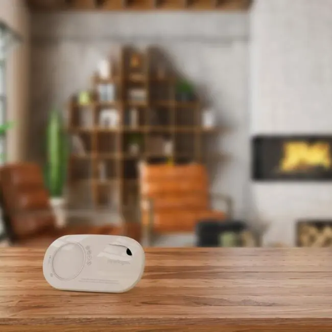 An FA3328-EUT carbon monoxide detector sits on a wooden table in a cosy living room.