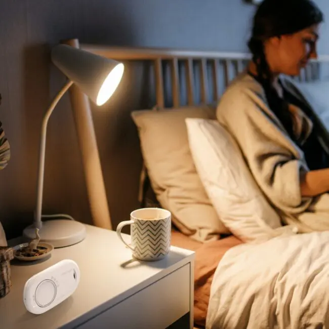 A woman sits on a bed near an FA3820-EUX10 bedside table holding a lamp, mug, and device.