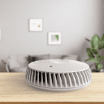 The FA6615-R smoke detector sits on a wooden table in a modern living room.