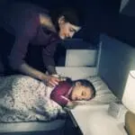 A woman tucks a child into bed beside a glowing FP1820W2-R baby monitor at night.