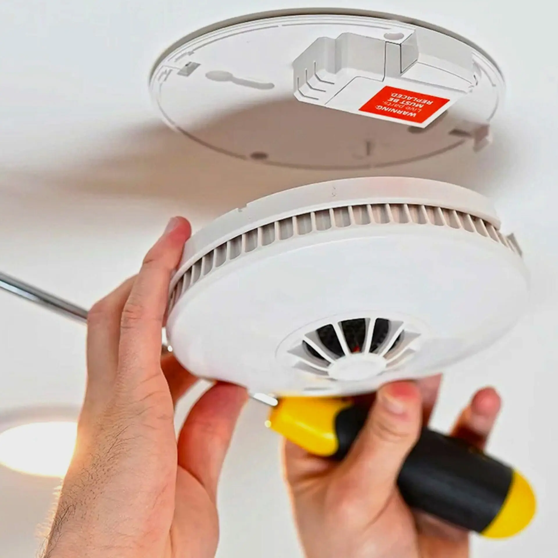 Hands mounting an HM-SN-1 smoke detector on the ceiling using a screwdriver.