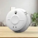 The SW1-PF-T white smoke detector sits on a wooden table with a blurred lounge behind.