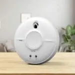 The SW1-R smoke detector sits on a wooden table with a blurred lounge background.