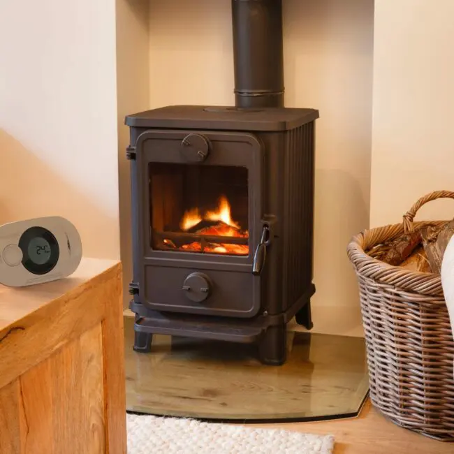 A FA3322-EUX10 wood-burning stove glows beside a log basket and a digital thermostatic controller.
