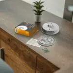 FA6720-R smoke detector with box, manual, and batteries on a wooden cupboard by a small plant.