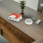 SB1-R smoke detector with box, batteries, and manual placed on wood beside a potted plant.