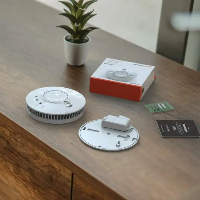 FP1640W2-R smoke detector and installation kit on a wooden surface beside a small plant.