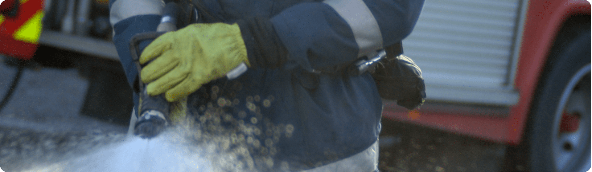 Firefighter in yellow gloves spraying water, promoting fire safety compliance, fire engine behind.