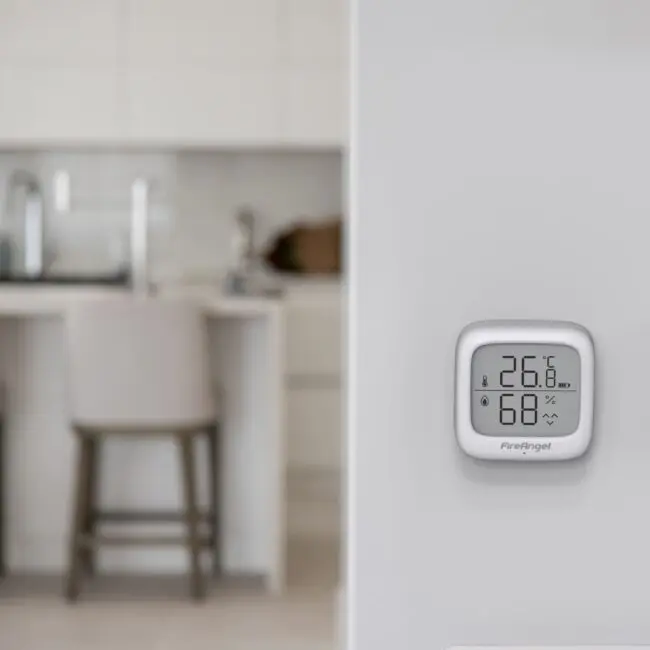 The FA7312-EUX4 digital thermostat is mounted on a white wall, kitchen and stools in the background.