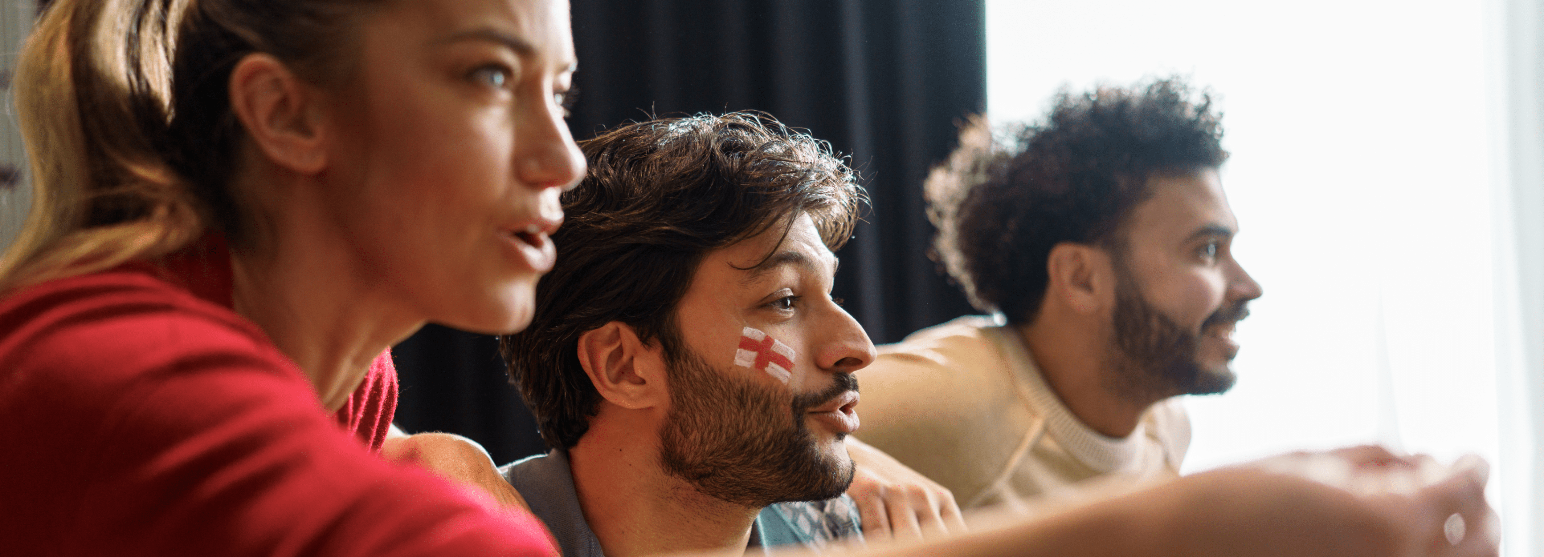 Three excited friends watch at home, one with an England flag on his cheek, hosting games safely.