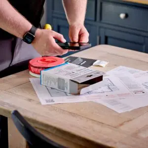 Person scanning a Spec+ Multi-Sensor Smoke, Heat & CO Alarm box with mobile on cluttered table.