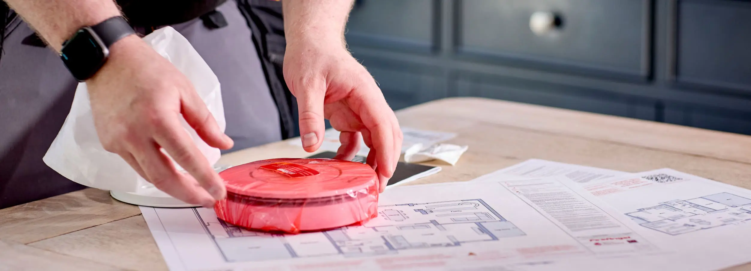 Hands placing red-wrapped object on blueprints, symbolising compliance support, atop wooden table.