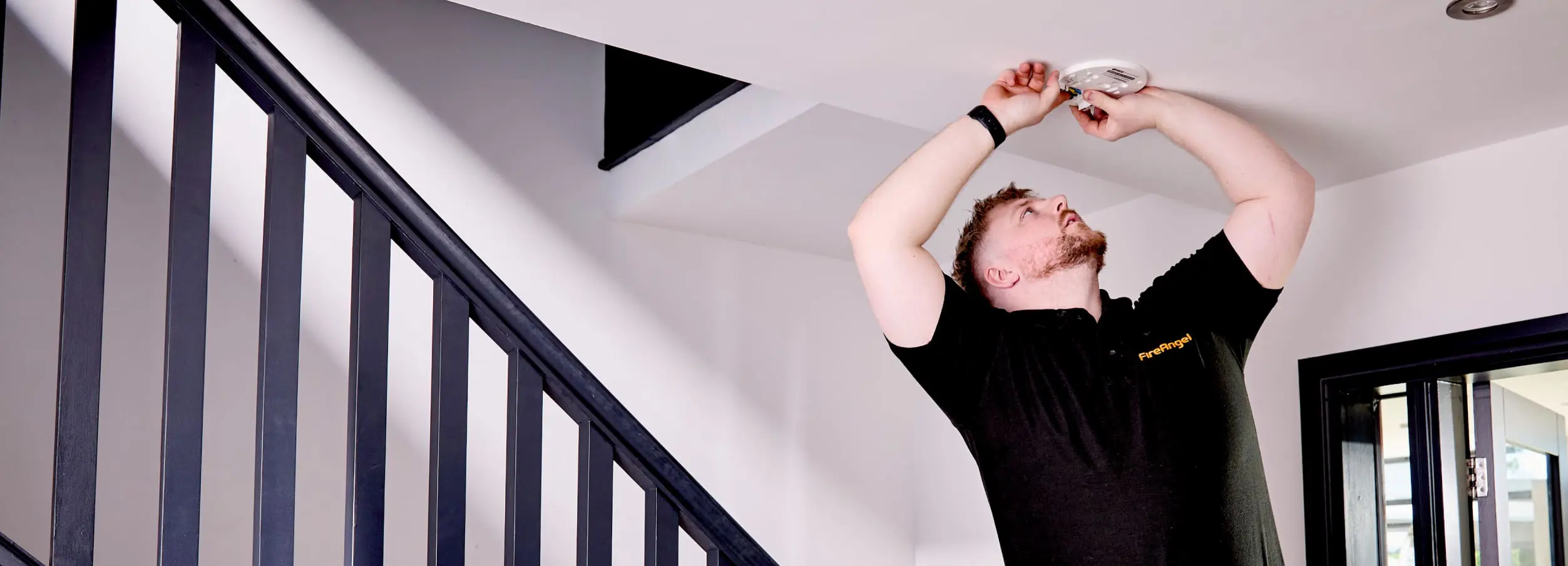 Man in black shirt installing a smoke detector for compliance support near a white stairway ceiling.