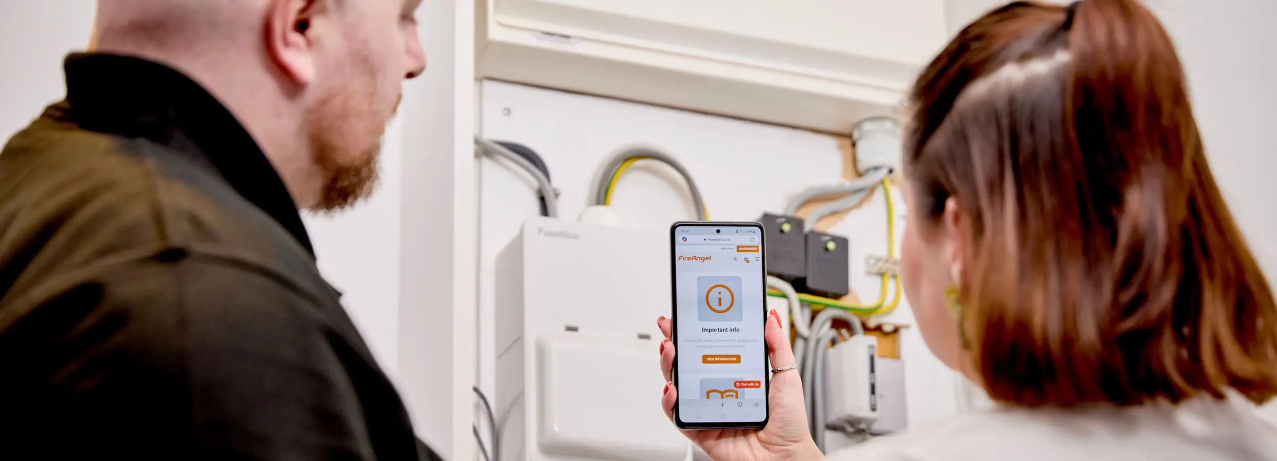 Two people check a mobile app for compliance support near electrical equipment on a wall.