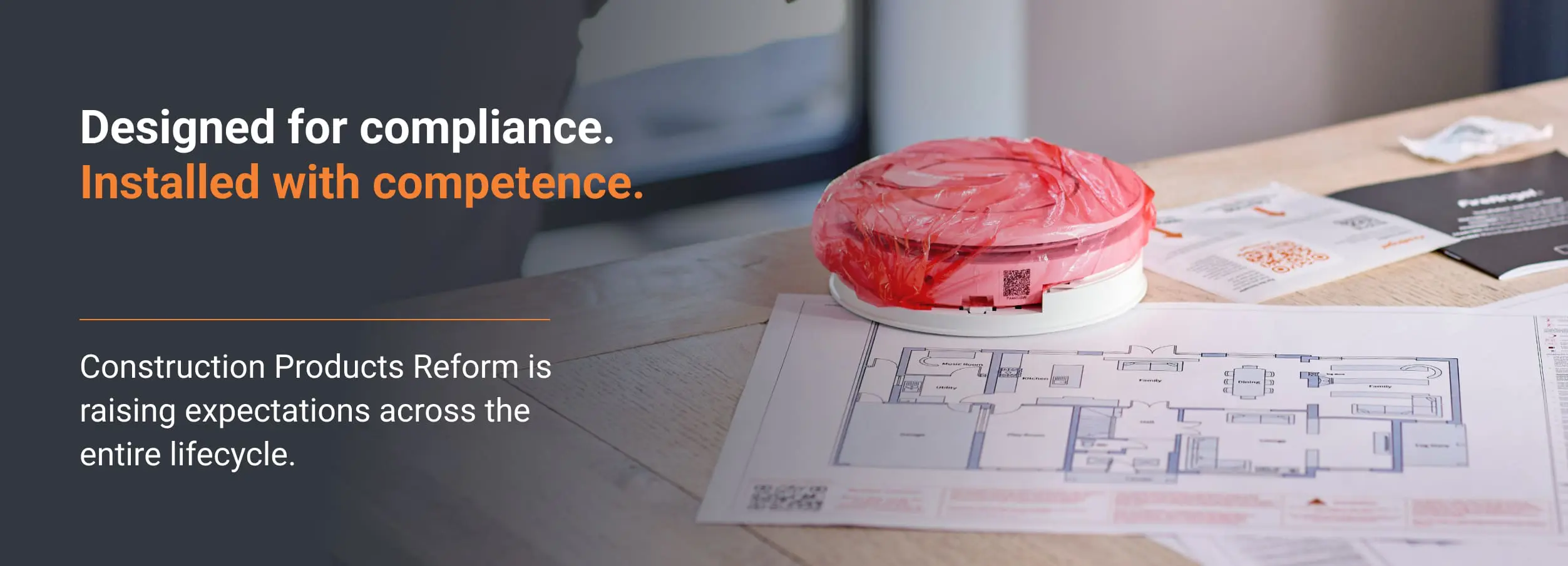 Round smoke alarm, key for compliance, sits on table beside blueprints and leaflets.