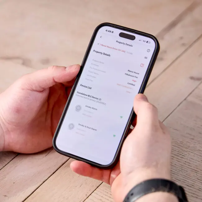 Hands holding a mobile showing Angel Pro App property details and connected alarm devices.